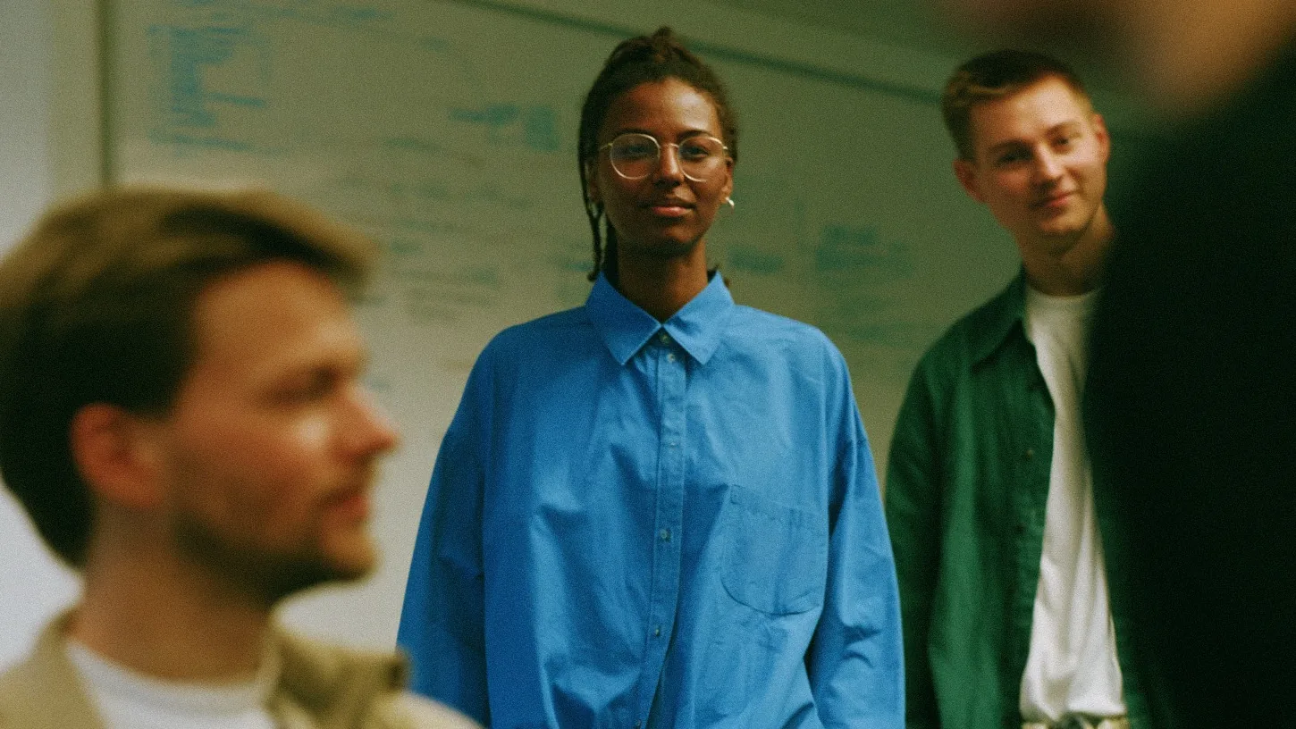 Three colleagues in a studio, a woman in a blue shirt and round glasses at the center
